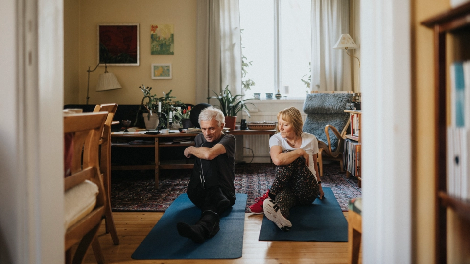 Bilden visar två äldre personer, en man och en kvinna, som sitter på varsin träningsmatta på golvet i ett mysigt vardagsrum. De gör en stretchövning och ser avslappnade ut. Kvinnan tittar på mannen och ler. Rummet är hemtrevligt inrett med träbord och stolar, en soffa, många växter och tavlor på väggarna.